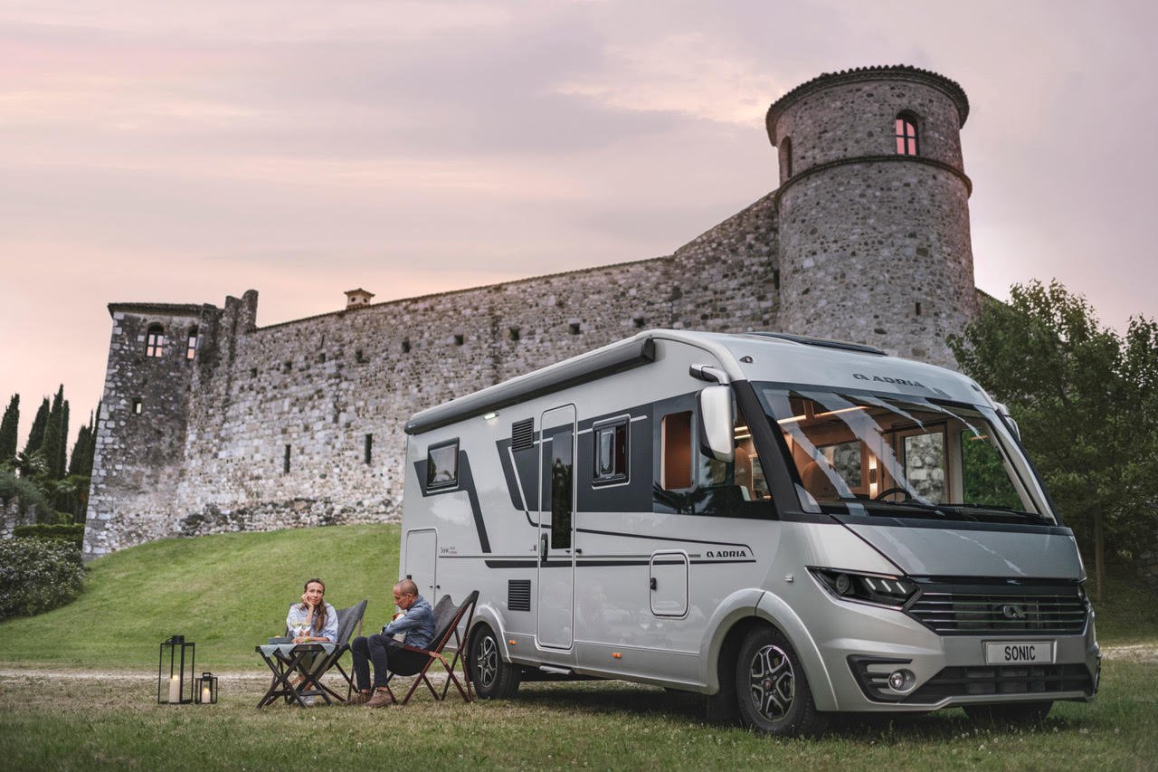 Modern husbil parkerad framför ett gammalt stenfort där två personer sitter utomhus vid campingplatsen.
